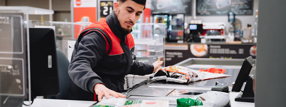 Eine Person in Selgros-Arbeitskleidung arbeitet an einer Kasse, scannt Waren, während im Hintergrund weitere Bereiche der Filiale zu sehen sind.