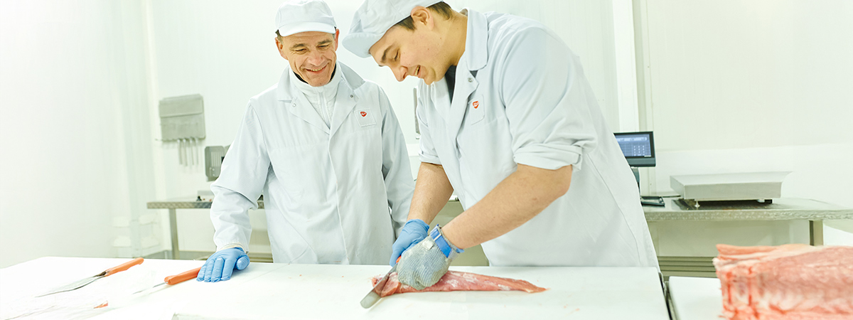 Zwei Personen stehen in weißer Arbeitskleidung in einer industriellen Umgebung. Eine Person schneidet ein Stück Fleisch.