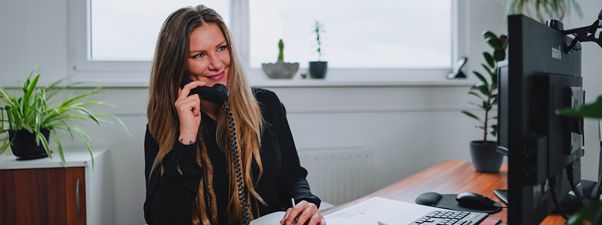 Eine Person sitzt an einem Schreibtisch in einem Büro, hält ein Telefon und scheint in ein Gespräch vertieft zu sein, während im Hintergrund Pflanzen und Fenster zu sehen sind.